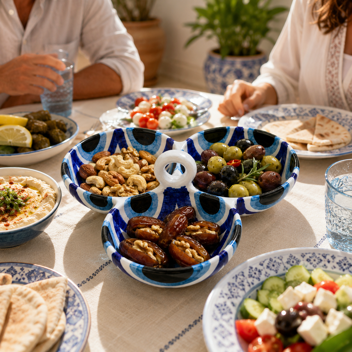 A three-compartment ceramic serving bowl with a blue and white eye pattern, filled with dates, nuts, and olives, set on a table with other Mediterranean dishes and people dining in the background.