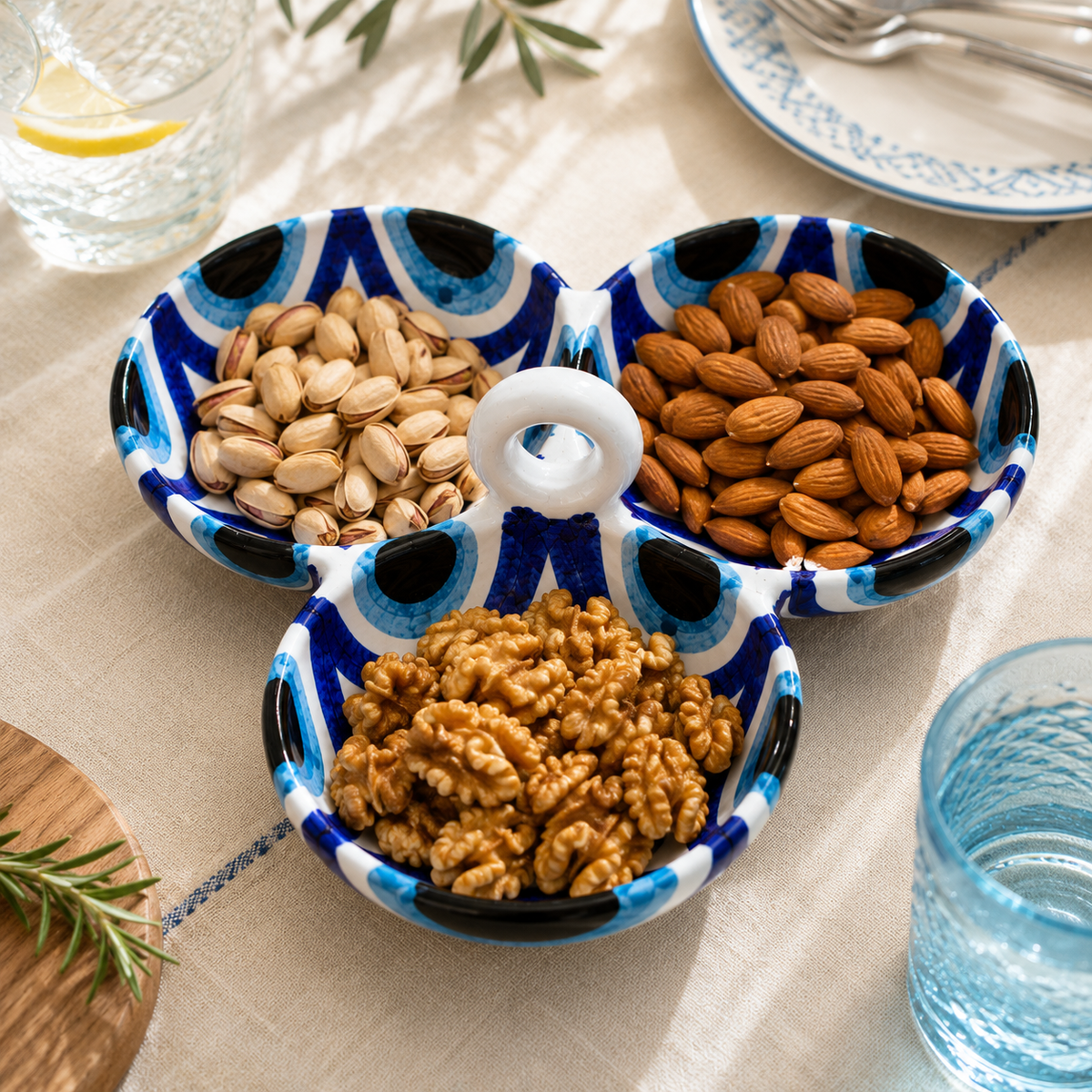 Three small ceramic serving bowls with a blue and black eye-like pattern and a white ring handle, arranged on a beige tablecloth and filled with pistachios, almonds, and walnuts.