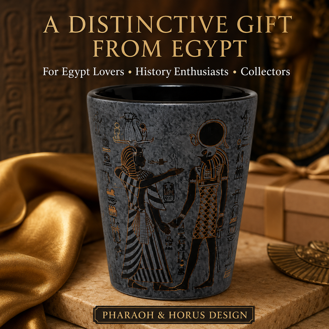 A dark gray ceramic mug with gold Egyptian hieroglyphic designs of Pharaoh and Horus, displayed on a stone pedestal with gold fabric and a wrapped gift box in the background.