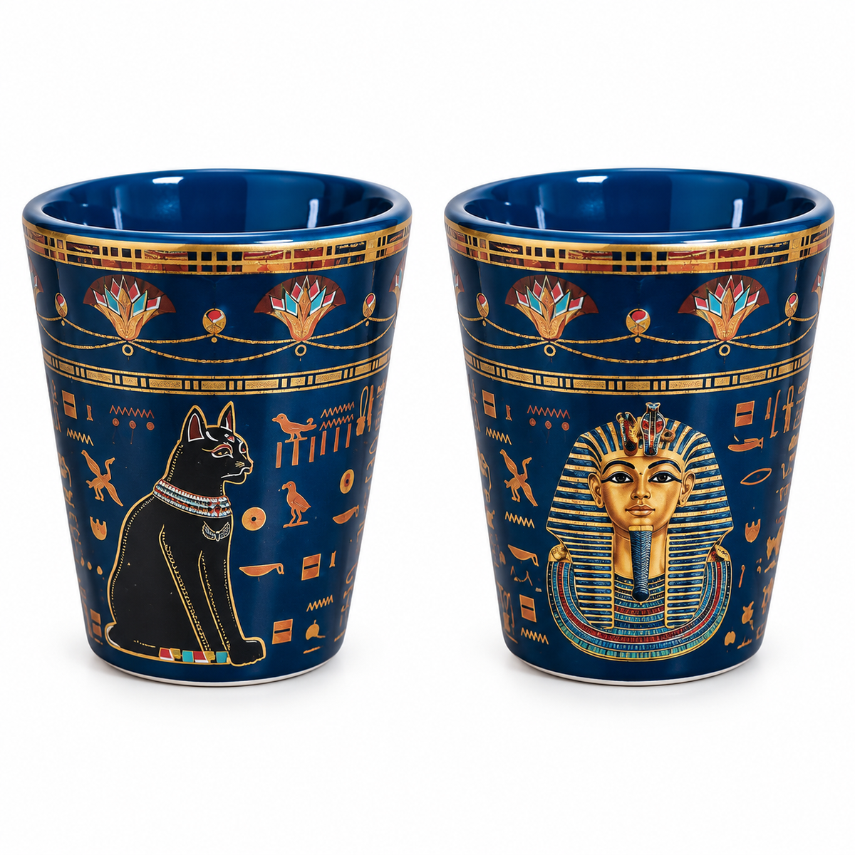 Two blue ceramic shot glasses with gold and multicolor Egyptian-themed designs, one featuring a black cat and the other a gold pharaoh mask, on a white background.