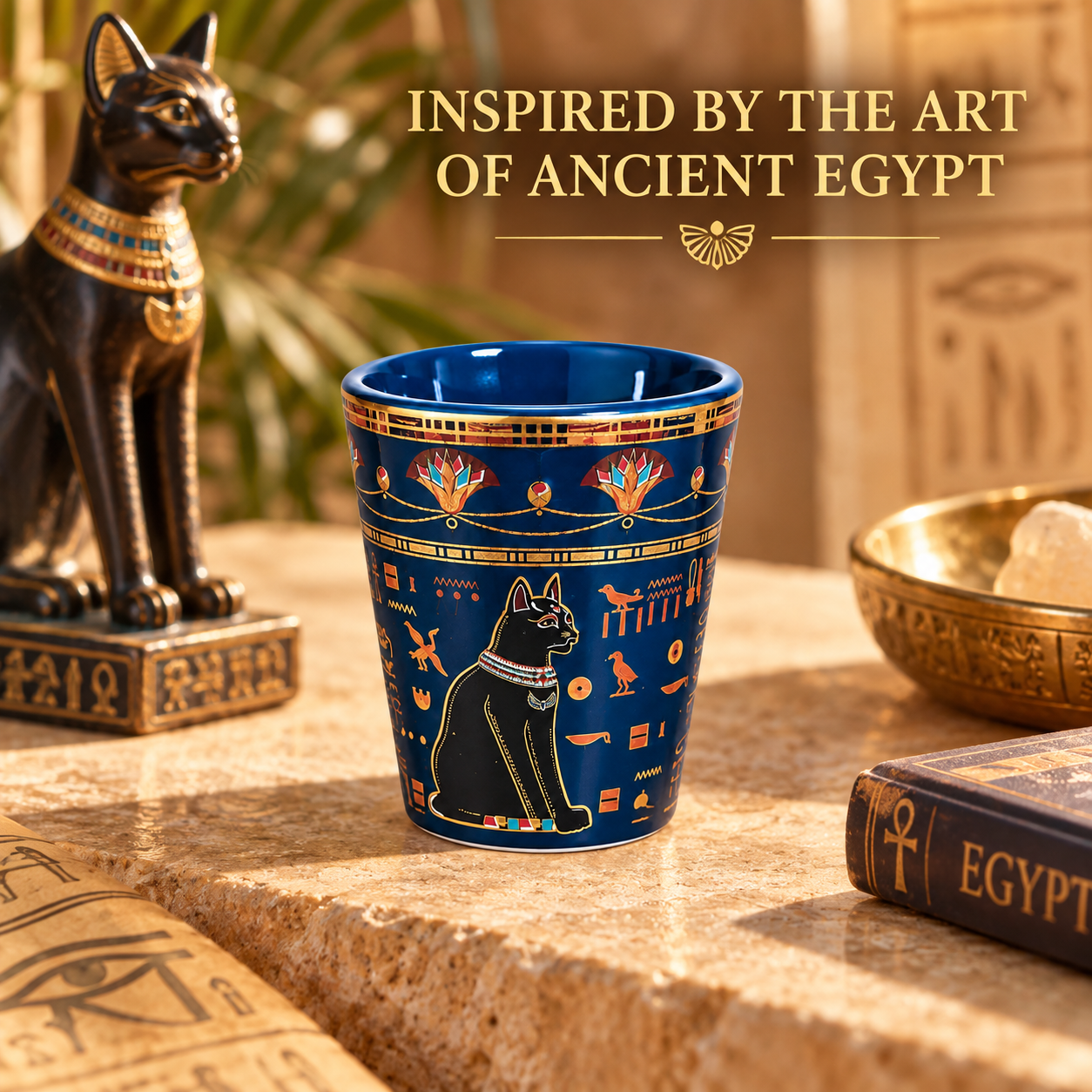 A dark blue ceramic cup with ancient Egyptian motifs, including a black cat and gold hieroglyphs, displayed on a stone surface next to an Egyptian statue and a book, with text reading 'INSPIRED BY THE ART OF ANCIENT EGYPT'.