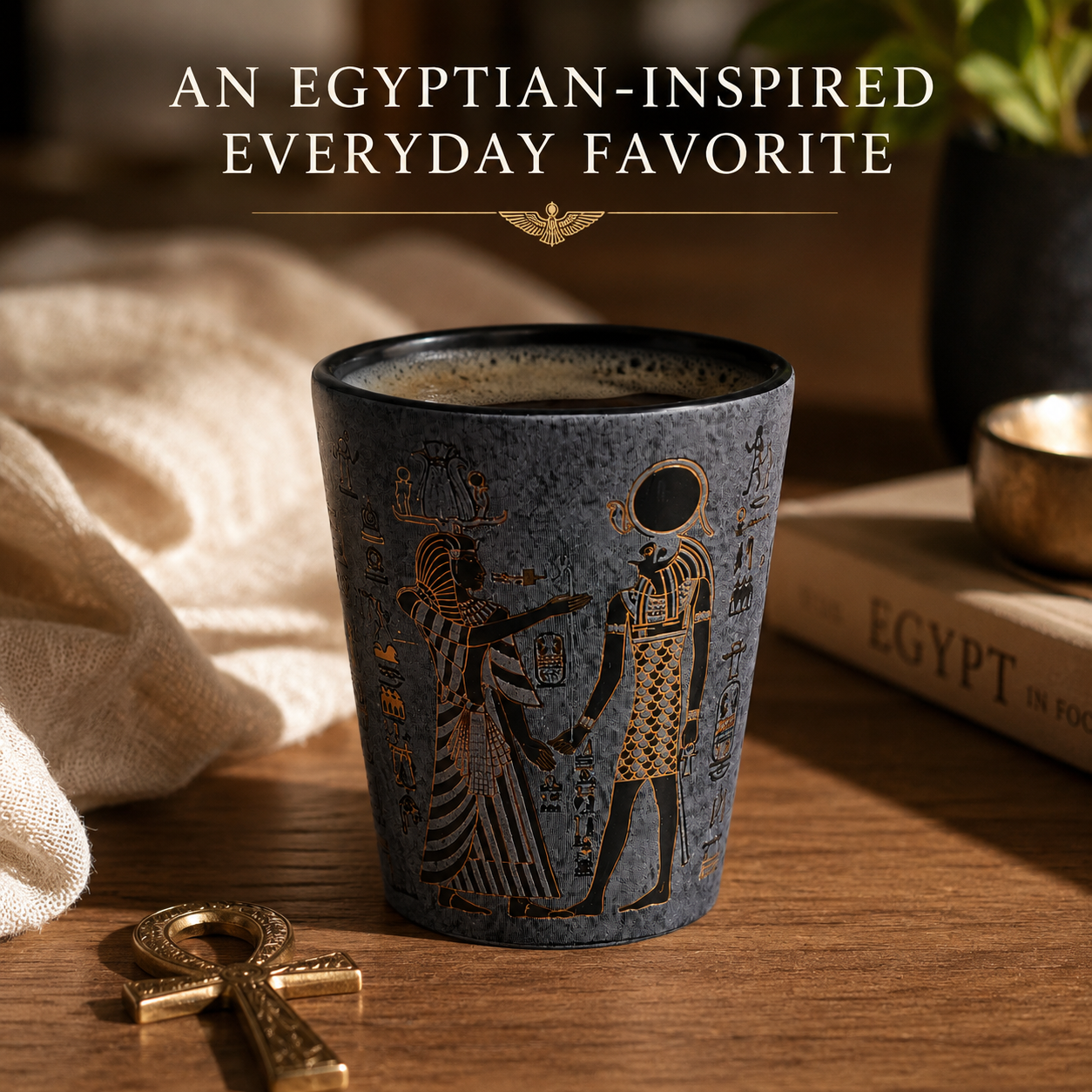 A dark blue-gray ceramic cup with gold Egyptian-style artwork and hieroglyphs, set on a wooden table with an ankh symbol, draped fabric, and a book in the background. Text at the top reads 'AN EGYPTIAN-INSPIRED EVERYDAY FAVORITE'.