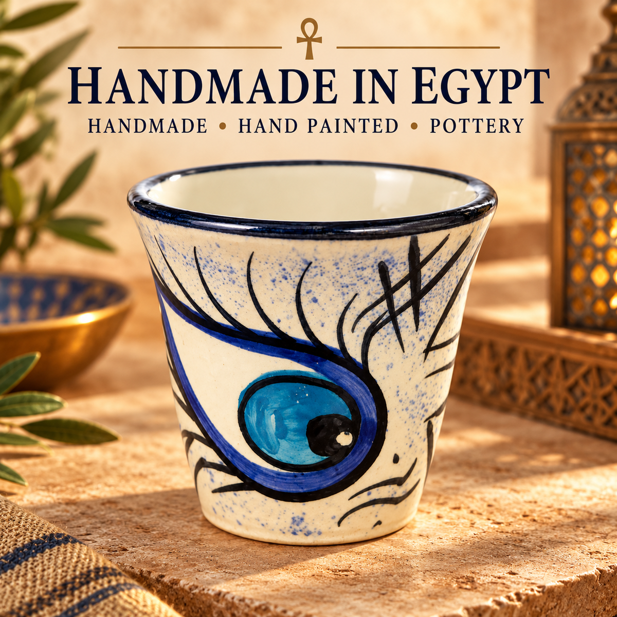 A handmade ceramic cup with a blue and black painted eye design on a cream background, set on a wooden surface with decorative props. Text at the top reads 'HANDMADE IN EGYPT' and 'HANDMADE • HAND PAINTED • POTTERY'.