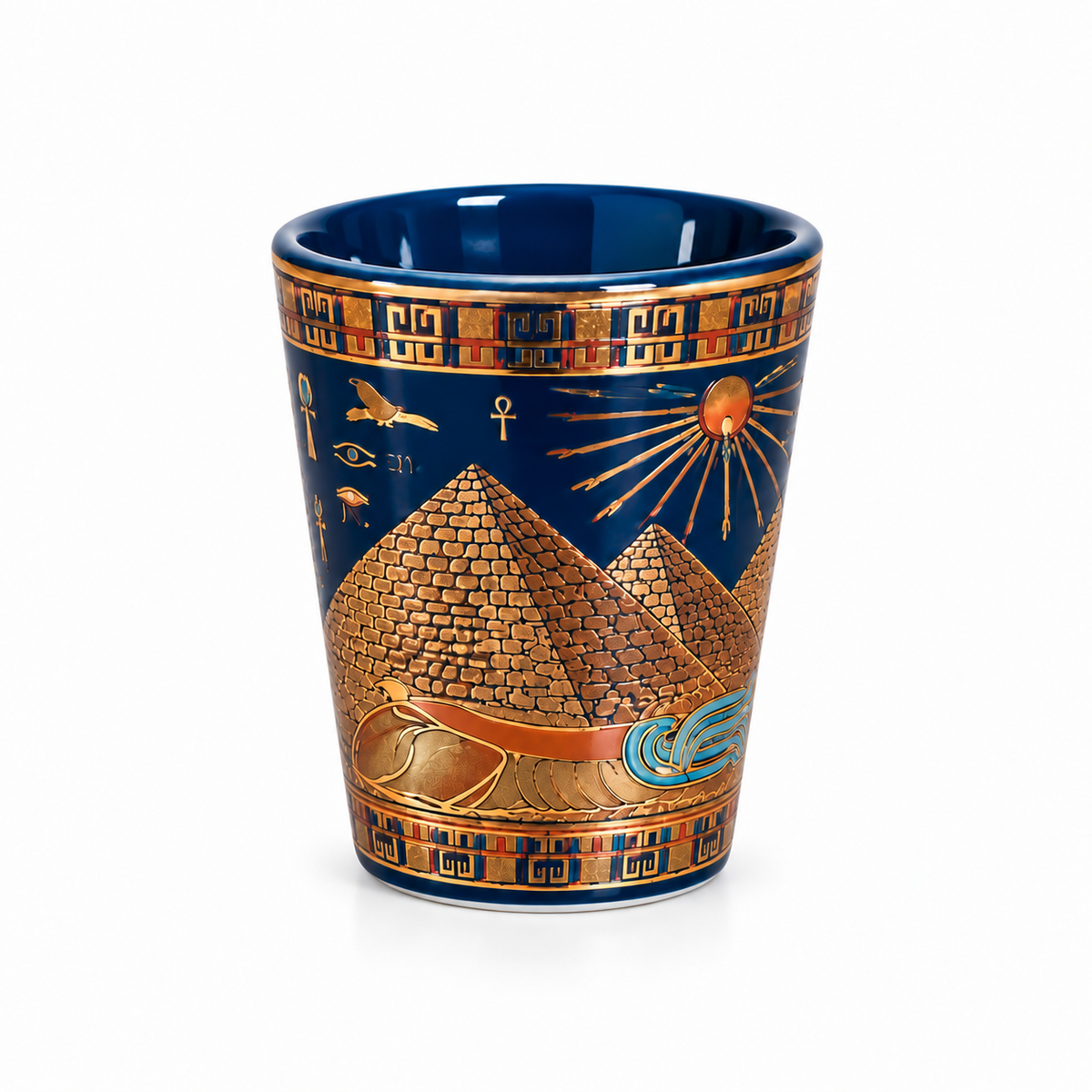 A ceramic shot glass featuring an Egyptian theme with gold pyramids, a sun, and hieroglyphs on a dark blue background, set against a plain white studio backdrop.