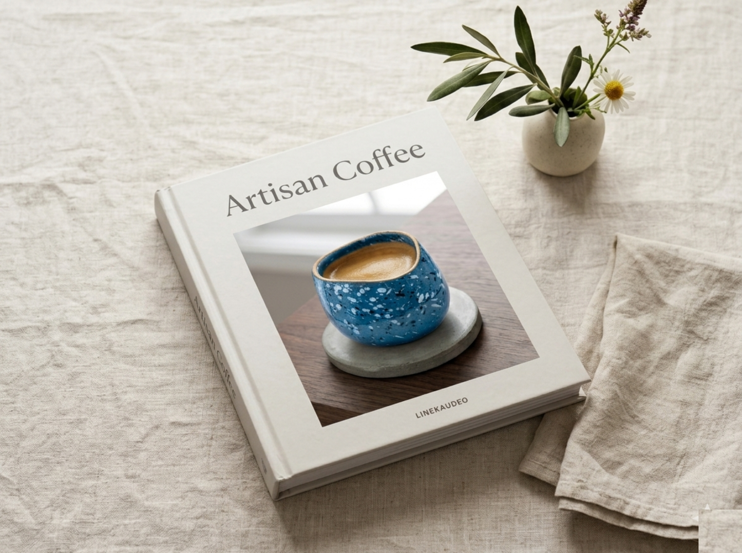 Book titled 'Artisan Coffee' with a blue coffee cup on the cover, placed on a light surface with a plant and napkin.