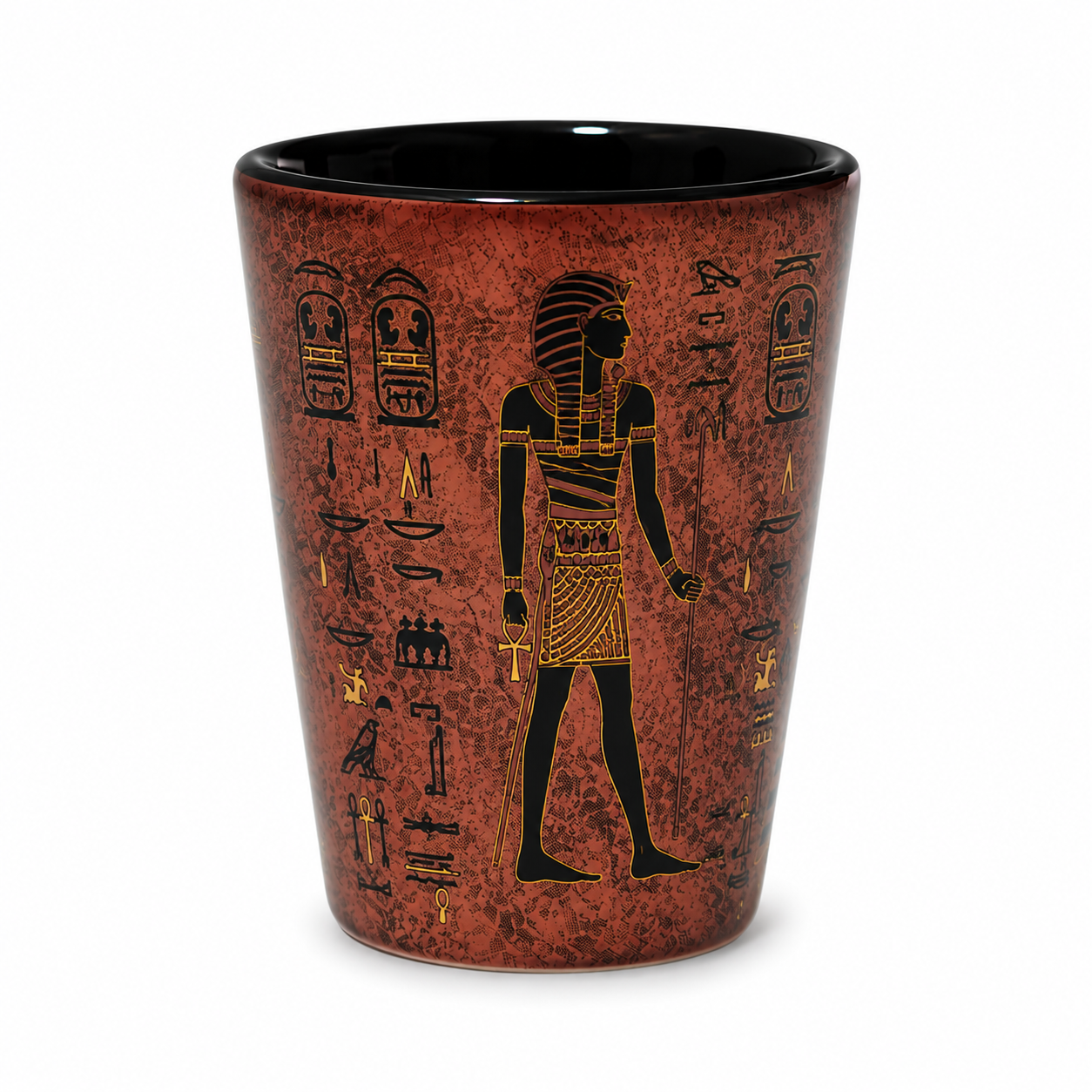 A tapered shot glass featuring an ancient Egyptian design with a textured reddish-brown body, black hieroglyphs, and a black pharaoh silhouette, set against a plain white background.