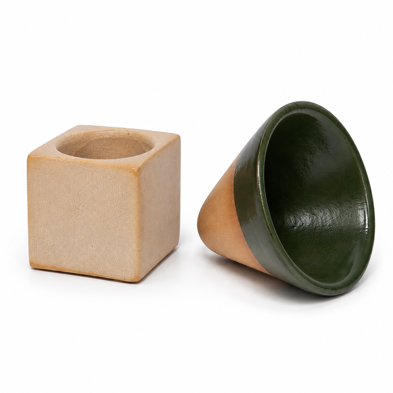 Two ceramic tableware pieces on a white background: a matte beige square cup with a circular opening, and a glossy dark green cone-shaped cup lying on its side.