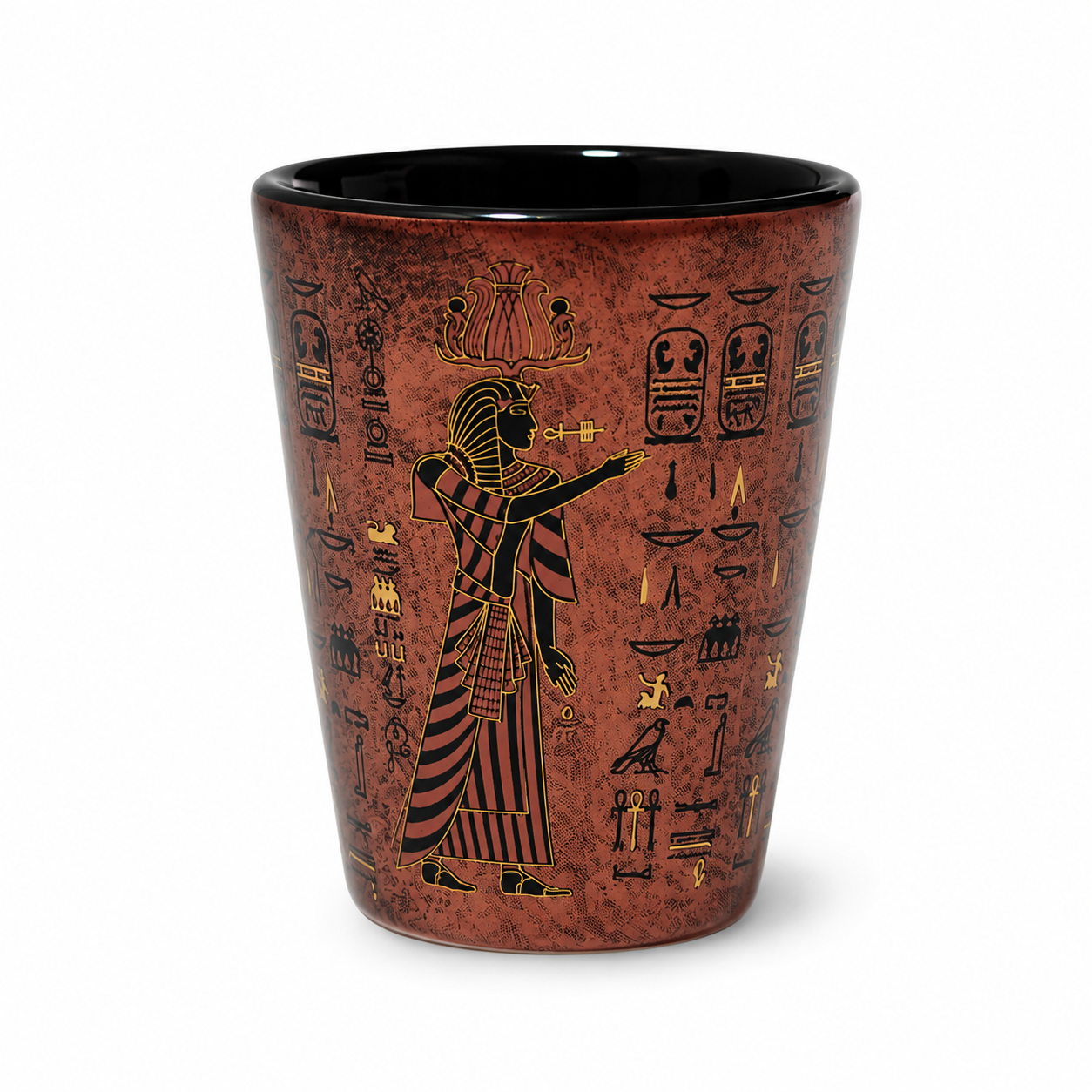 A tapered shot glass featuring an ancient Egyptian design with a profile figure, hieroglyphs, and cartouches in gold and black on a textured reddish-brown background, set against a plain white background.