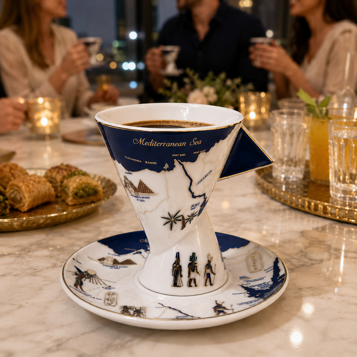 A ceramic pour-over coffee cup and saucer set with a Mediterranean Sea map design, styled on a marble table in a restaurant setting with people holding drinks in the background.