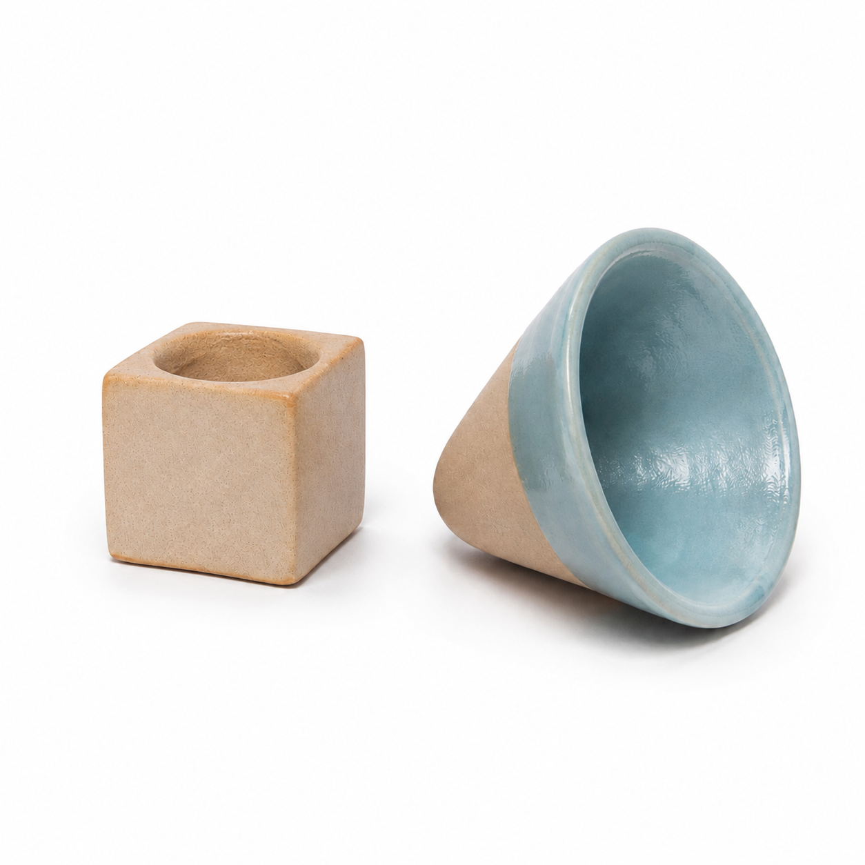 Two small ceramic objects on a white background: a matte tan cube with a circular hole at the top, and a matching cone-shaped cup lying on its side, showing a light blue glazed interior and tan exterior.