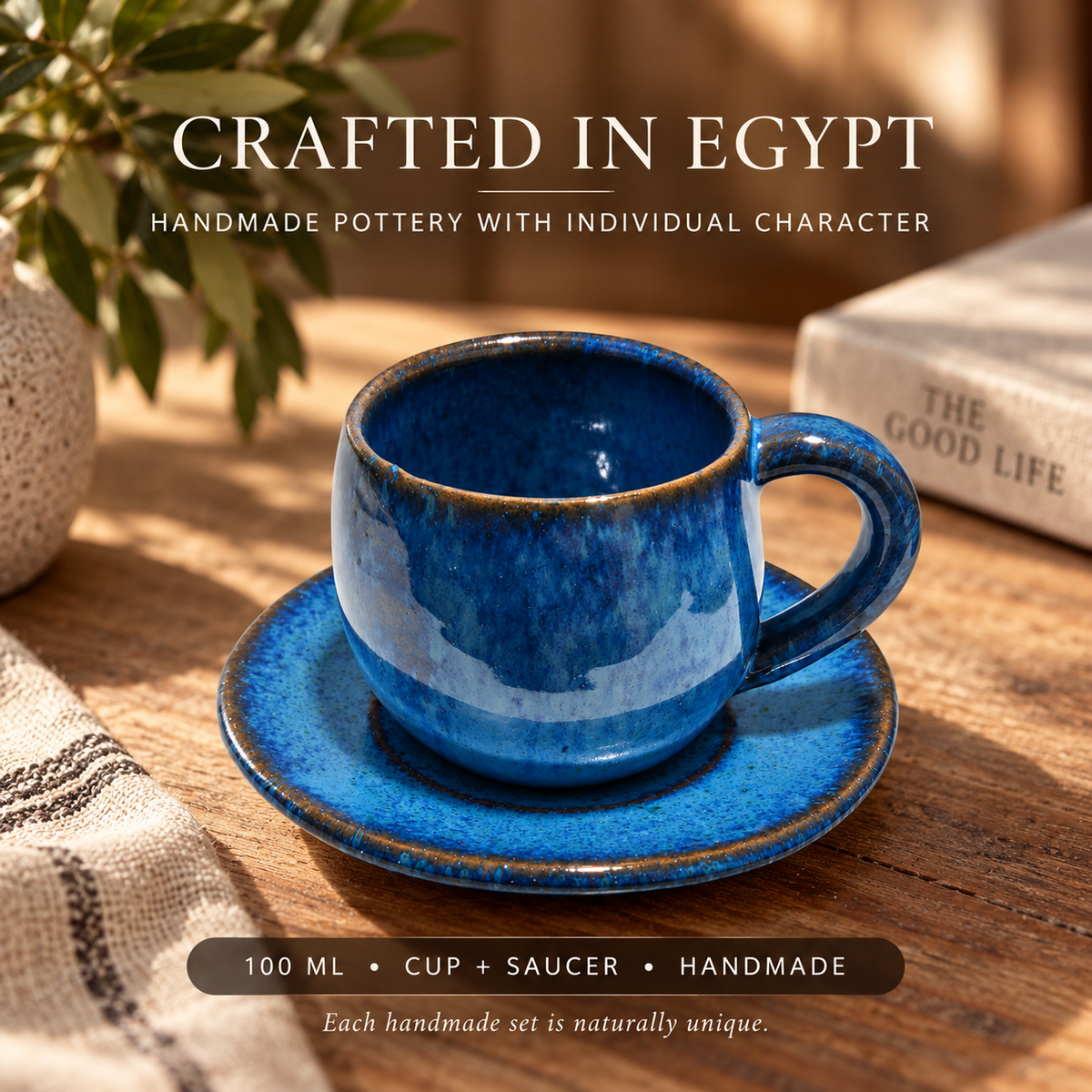 A blue glazed ceramic cup and saucer set on a wooden table, with decorative text reading 'CRAFTED IN EGYPT' and '100 ML • CUP + SAUCER • HANDMADE'. A plant, textured vase, and book are softly blurred in the background.