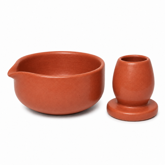 A terracotta-colored ceramic set featuring a shallow bowl with a pouring spout and a small rounded cup with a matching base, both photographed on a clean white background.