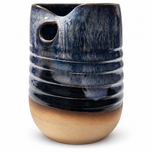 A handmade ceramic pitcher with a dark blue to beige gradient glaze, featuring a ribbed body, a small side hole, and a pouring spout, set against a plain white background.